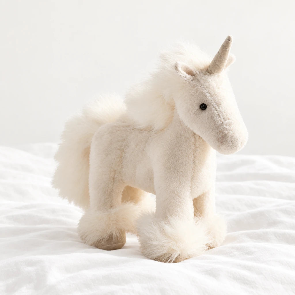Licorne en peluche debout à crinière colorée blanche posée sur un lit blanc, en pleine lumière