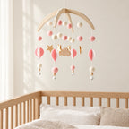 Mobile Bébé Bohème Rose en bois suspendu au-dessus d’un lit bébé en bois clair, dans une chambre douce