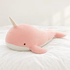 Peluche baleine magique géante rose pâle posée sur un lit blanc, en pleine lumière