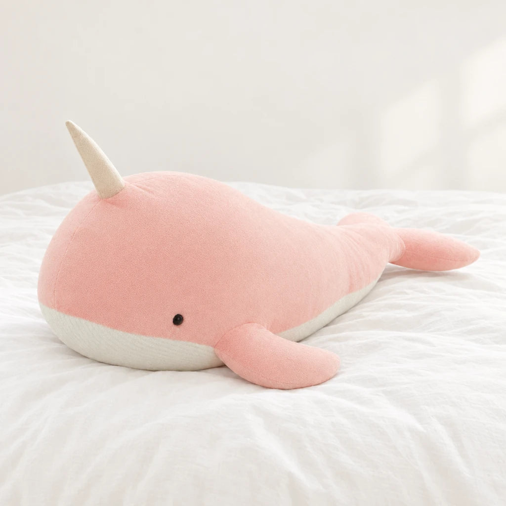 Peluche baleine magique géante rose pâle posée sur un lit blanc, en pleine lumière
