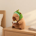 Peluche capybara avec boisson et bonnet rigolo modèle avocat posée sur une table de chevet en bois clair