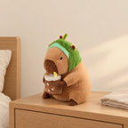 Peluche capybara avec boisson et bonnet rigolo modèle avocat posée sur une table de chevet en bois clair