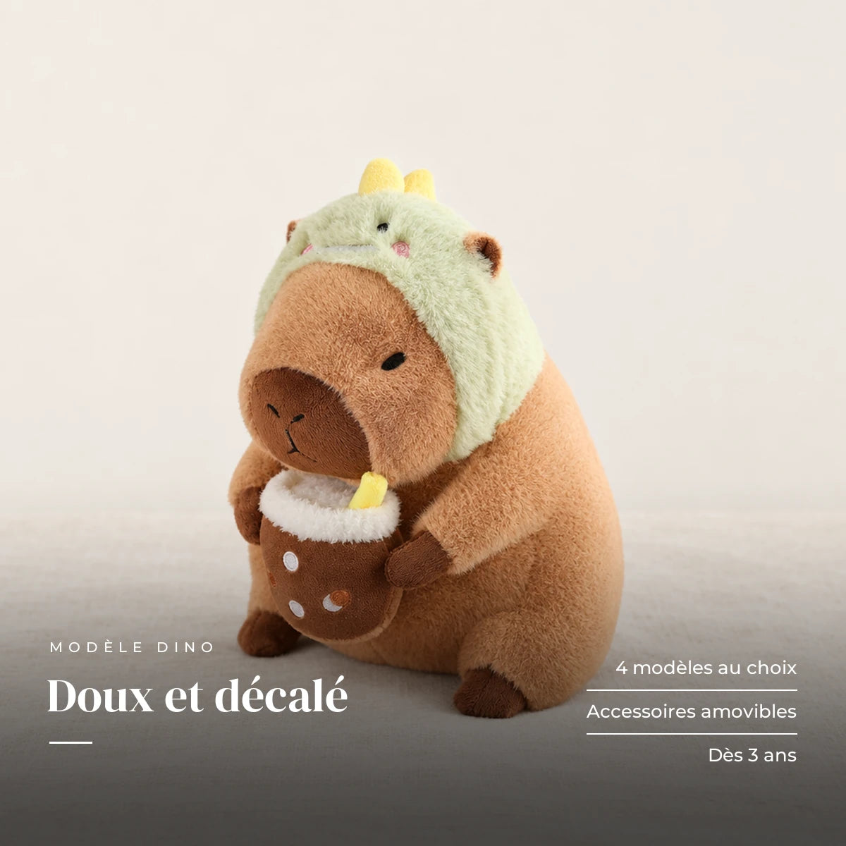 Peluche capybara avec boisson et bonnet rigolo modèle dinosaure sur fond crème, en studio. Doux et décalé : 4 modèles au choix, accessoires amovibles, dès 3 ans