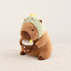 Peluche capybara avec boisson et bonnet rigolo modèle dinosaure sur fond crème, en studio