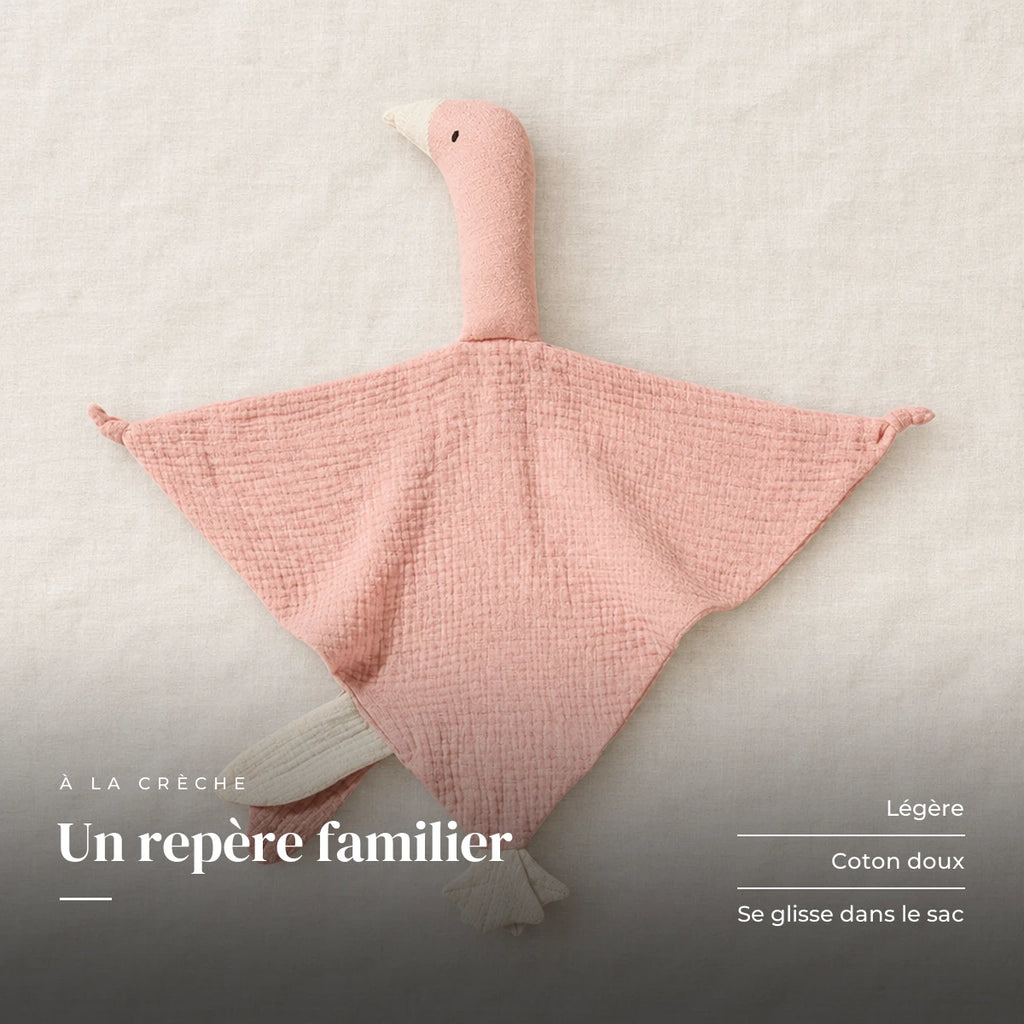 Peluche doudou oie pour bébé en coton rose présentée à plat sur du lin crème. Un repère familier : légère, coton doux, se glisse dans le sac