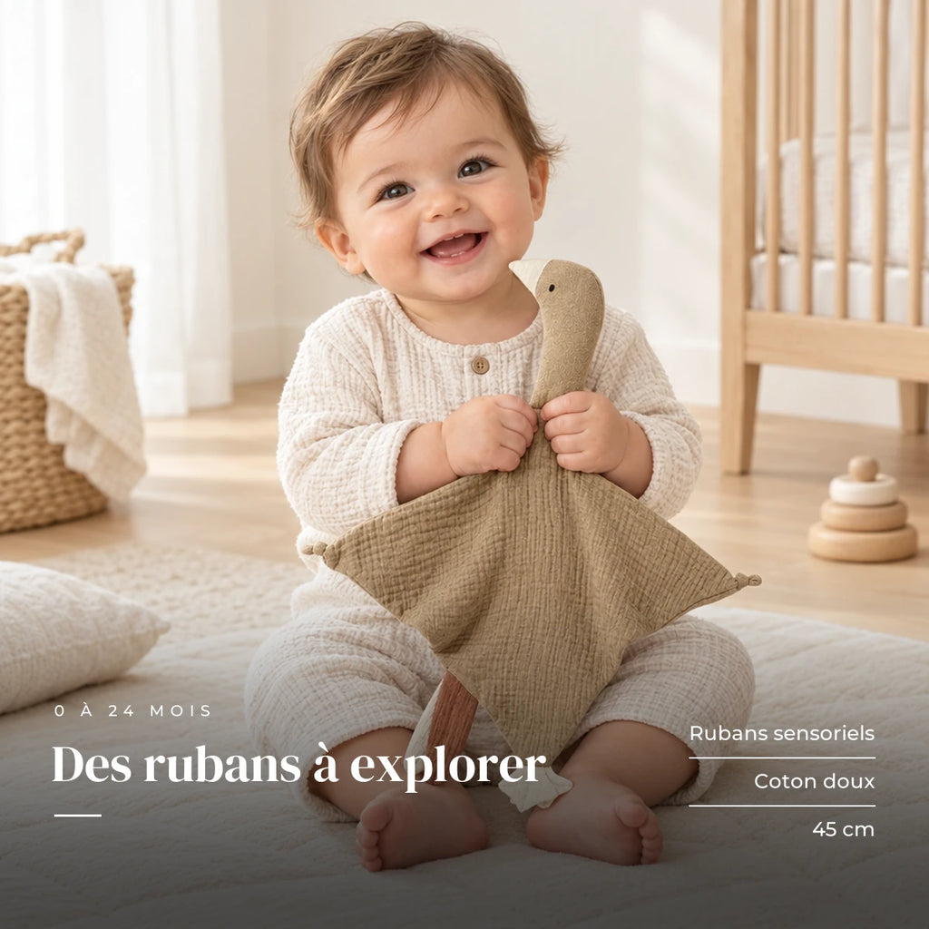Peluche doudou oie pour bébé en coton taupe dans les bras d’un bébé éveillé. Des rubans à explorer : rubans sensoriels, coton doux, 45 cm