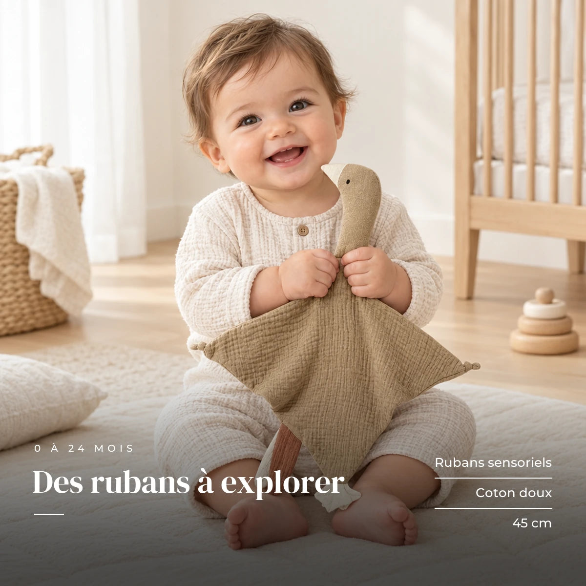 Peluche doudou oie pour bébé en coton taupe dans les bras d’un bébé éveillé. Des rubans à explorer : rubans sensoriels, coton doux, 45 cm