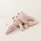Dragon en peluche aux couleurs pastel rose posé sur un lit en lin blanc