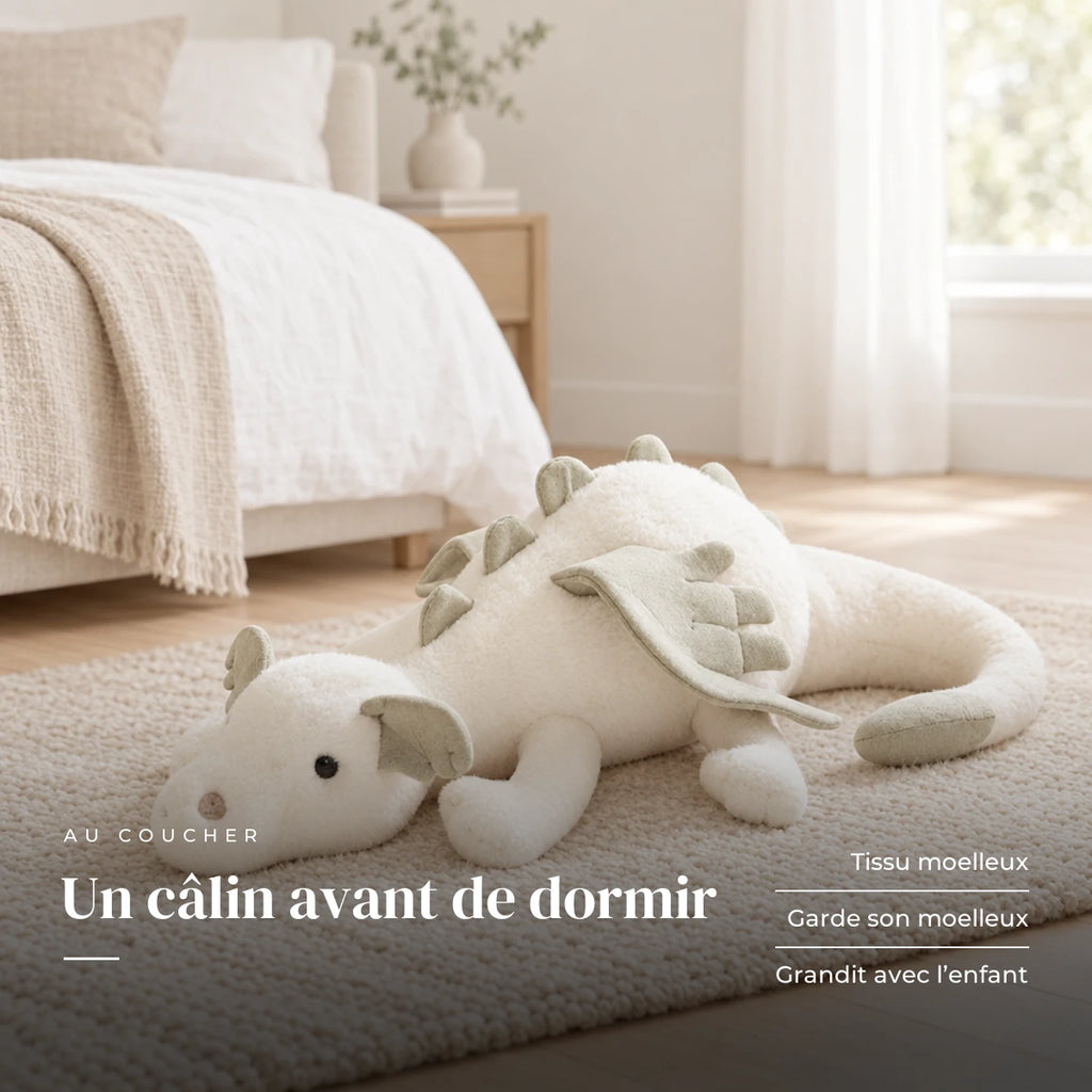 Dragon en peluche aux couleurs pastel posé sur un lit en lin blanc. Un câlin avant de dormir : tissu moelleux, garde son moelleux, grandit avec l’enfant