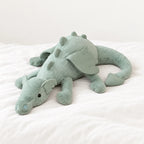 Dragon en peluche aux couleurs pastel vert posé sur un lit blanc, en pleine lumière