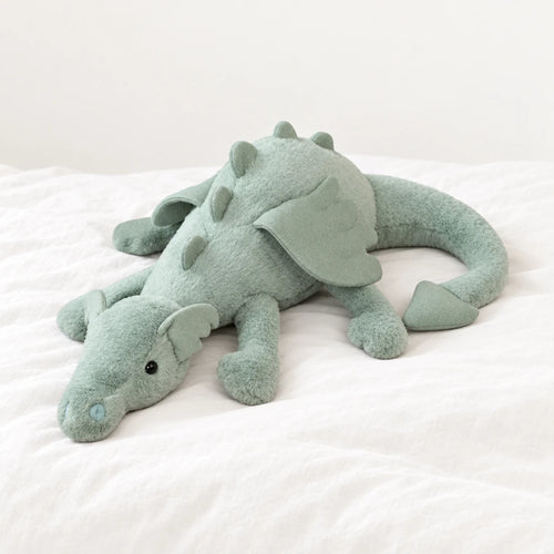 Dragon en peluche aux couleurs pastel vert posé sur un lit blanc, en pleine lumière