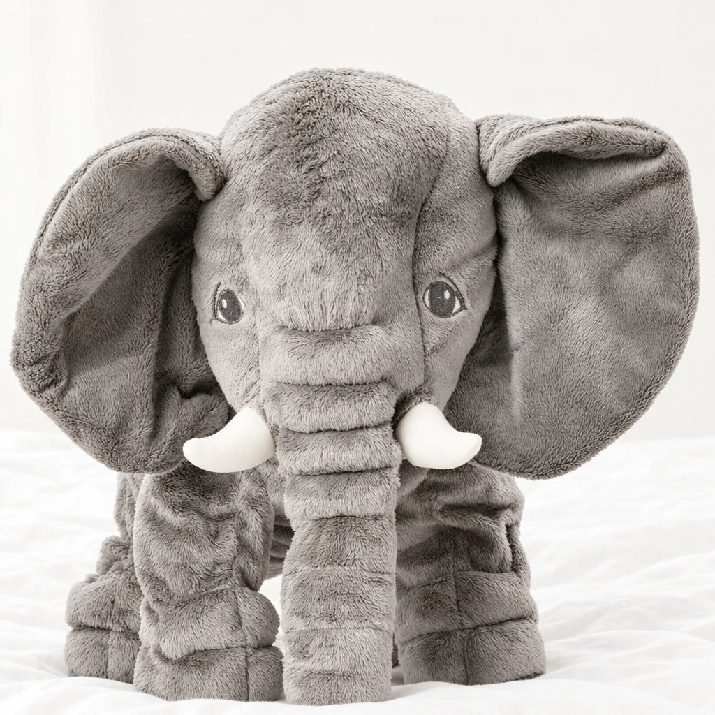 Peluche éléphant oreiller grise posée sur un lit blanc, en pleine lumière