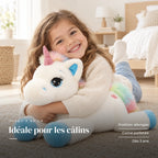 Peluche licorne arc-en-ciel allongée avec un enfant dans une chambre lumineuse. Idéale pour les câlins : position allongée, corne pailletée, dès 3 ans