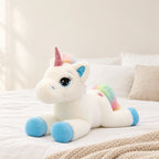 Peluche licorne arc-en-ciel allongée bleue posée sur un lit en lin blanc