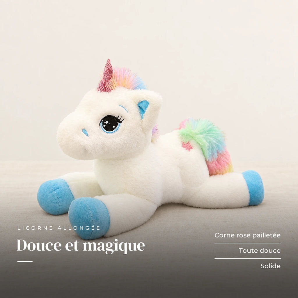 Peluche licorne arc-en-ciel allongée posée sur un lit en lin blanc. Douce et magique : corne rose pailletée, toute douce, solide