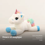 Peluche licorne arc-en-ciel allongée posée sur un lit en lin blanc. Douce et magique : corne rose pailletée, toute douce, solide