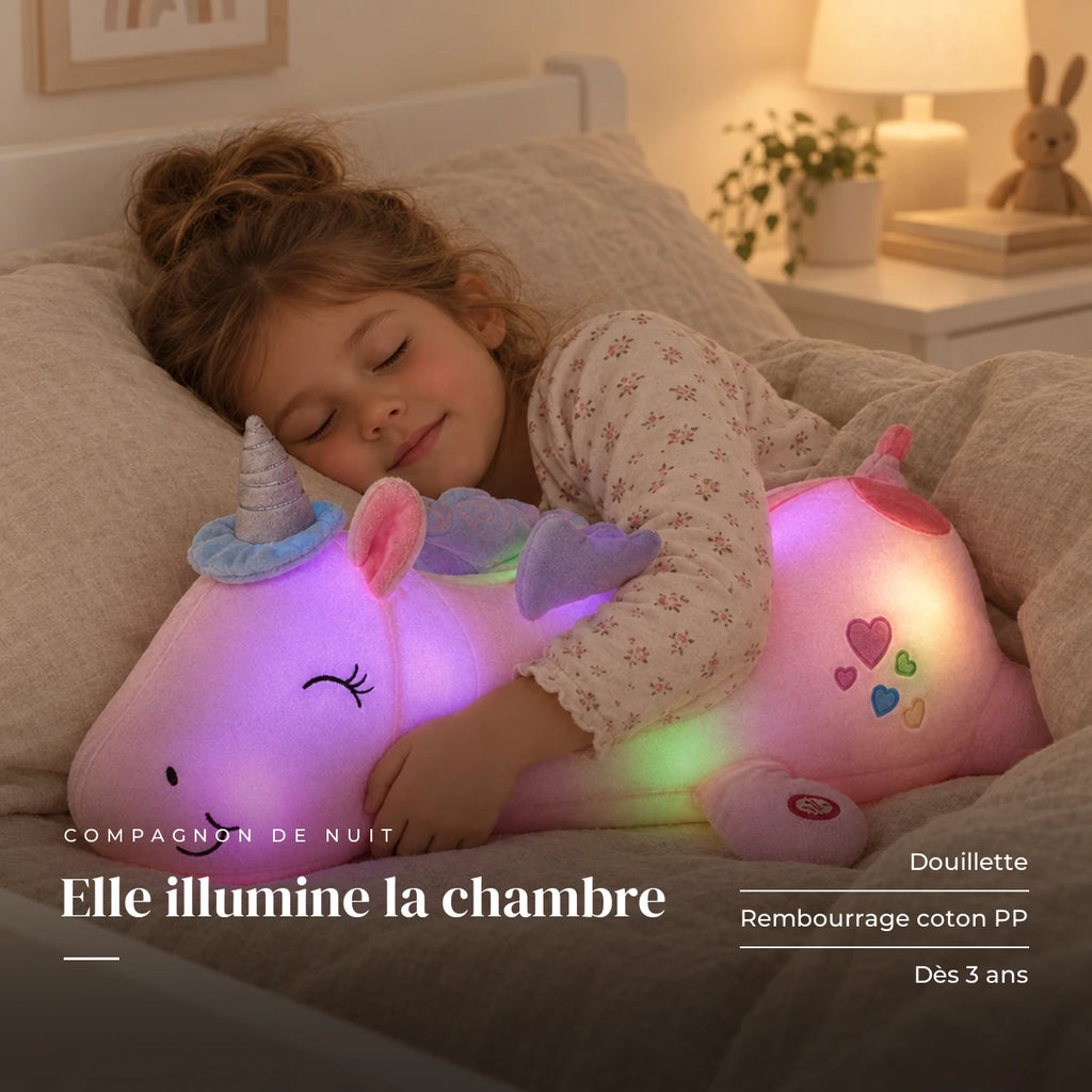 Peluche licorne rose veilleuse lumineuse et magique serrée par un enfant endormi dans son lit. Elle illumine la chambre : douillette, rembourrage coton PP, dès 3 ans