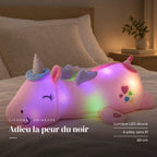 Peluche licorne rose veilleuse lumineuse et magique posée sur un lit en lin blanc. Adieu la peur du noir : lumière LED douce, à piles, sans fil, 60 cm