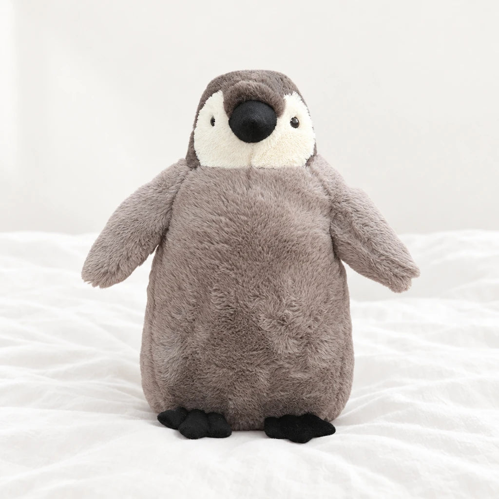 Peluche pingouin manchot câlin gris posée sur un lit blanc, en pleine lumière