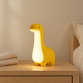 Veilleuse dinosaure bébé allumée sur une table de chevet en chêne dans une chambre d’enfant