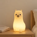 Veilleuse lama bébé rechargeable en silicone allumée sur une table de chevet en chêne dans une chambre d’enfant
