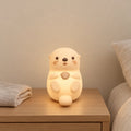 Veilleuse loutre en silicone rechargeable allumée sur une table de chevet en chêne dans une chambre d’enfant
