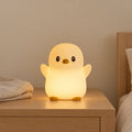 Veilleuse pingouin rechargeable allumée sur une table de chevet en chêne dans une chambre d’enfant